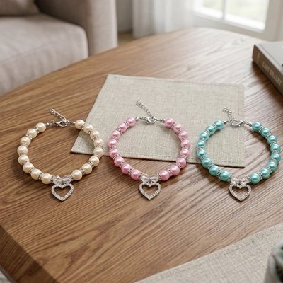 Trois colliers perles pour chat blanc rose et bleu avec pendentif cœur strass posés sur une table